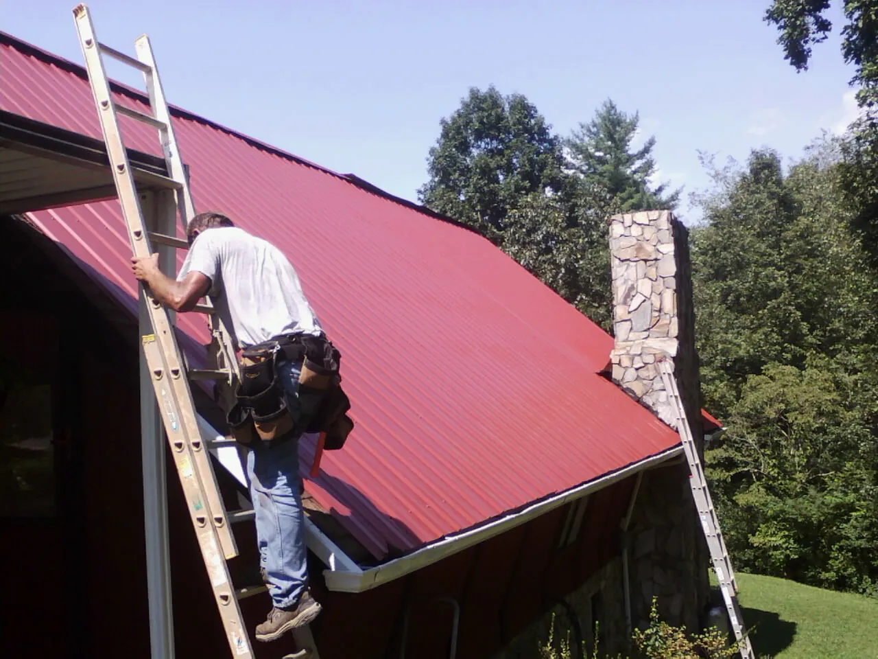Roofer on a ladder beside a red metal roof and stone chimney — Soffit & Fascia Repair by Roofing Brothers in Garrett